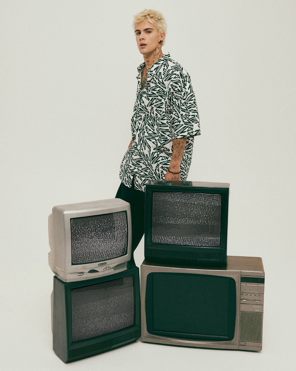 Young blond male model wearing a patterned white shirt standing behind a stack of vintage CRT television sets against a plain background