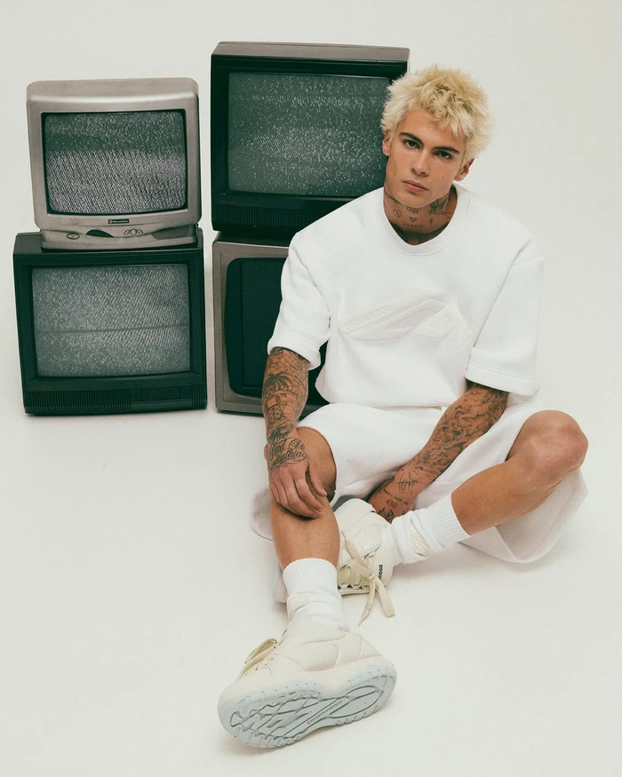 Blonde male model wearing white sweatshirt and shorts sitting next to stacked vintage televisions on a white background