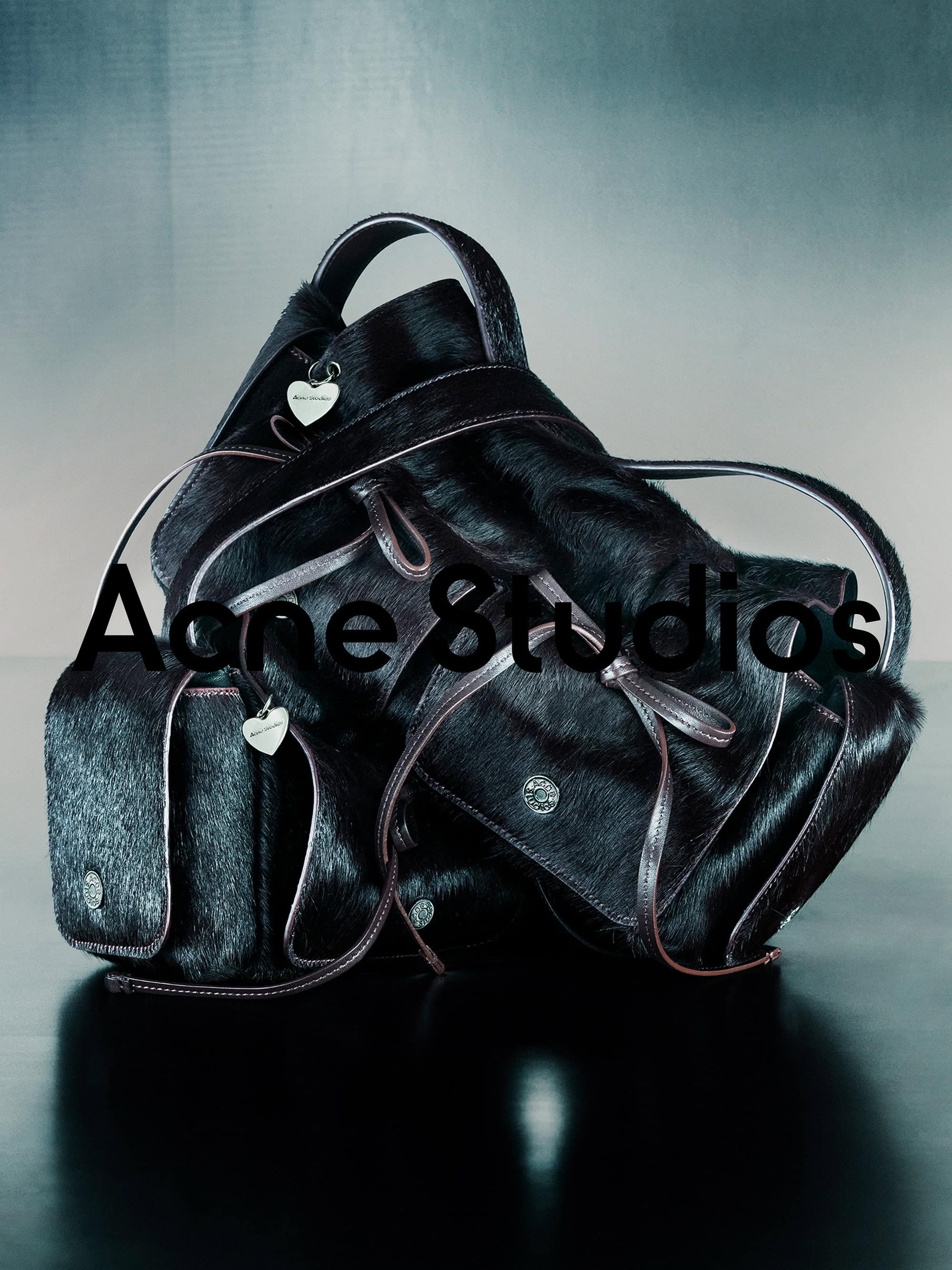 Stacked dark textured leather handbags with heart charms against a gradient background for Acne Studios