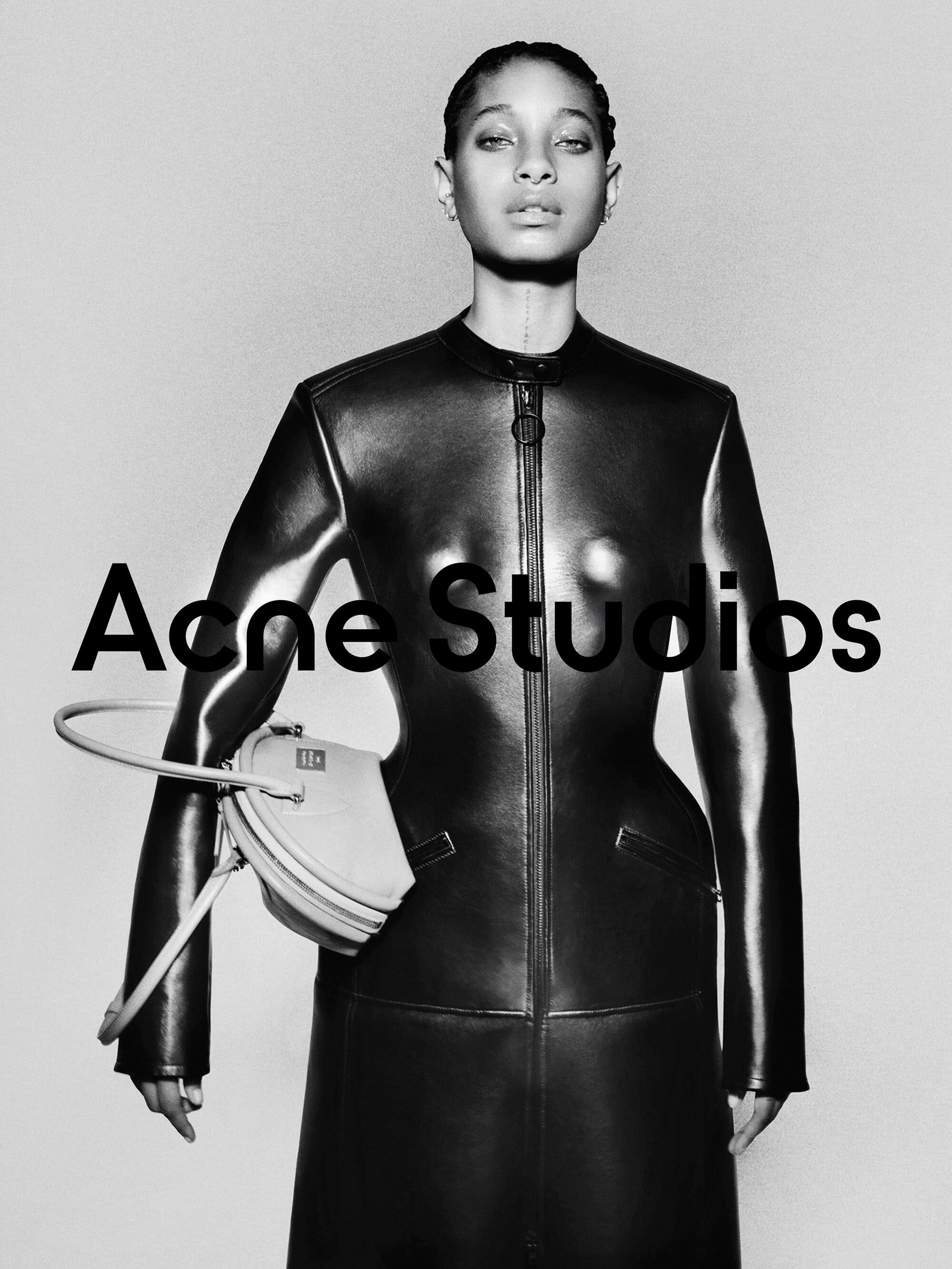 Willow Smith wearing a fitted black leather coat holding a handbag against a seamless grey backdrop for Acne Studios.