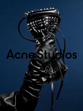 Gloved hand holding a black studded leather handbag against a solid dark blue background for Acne Studios