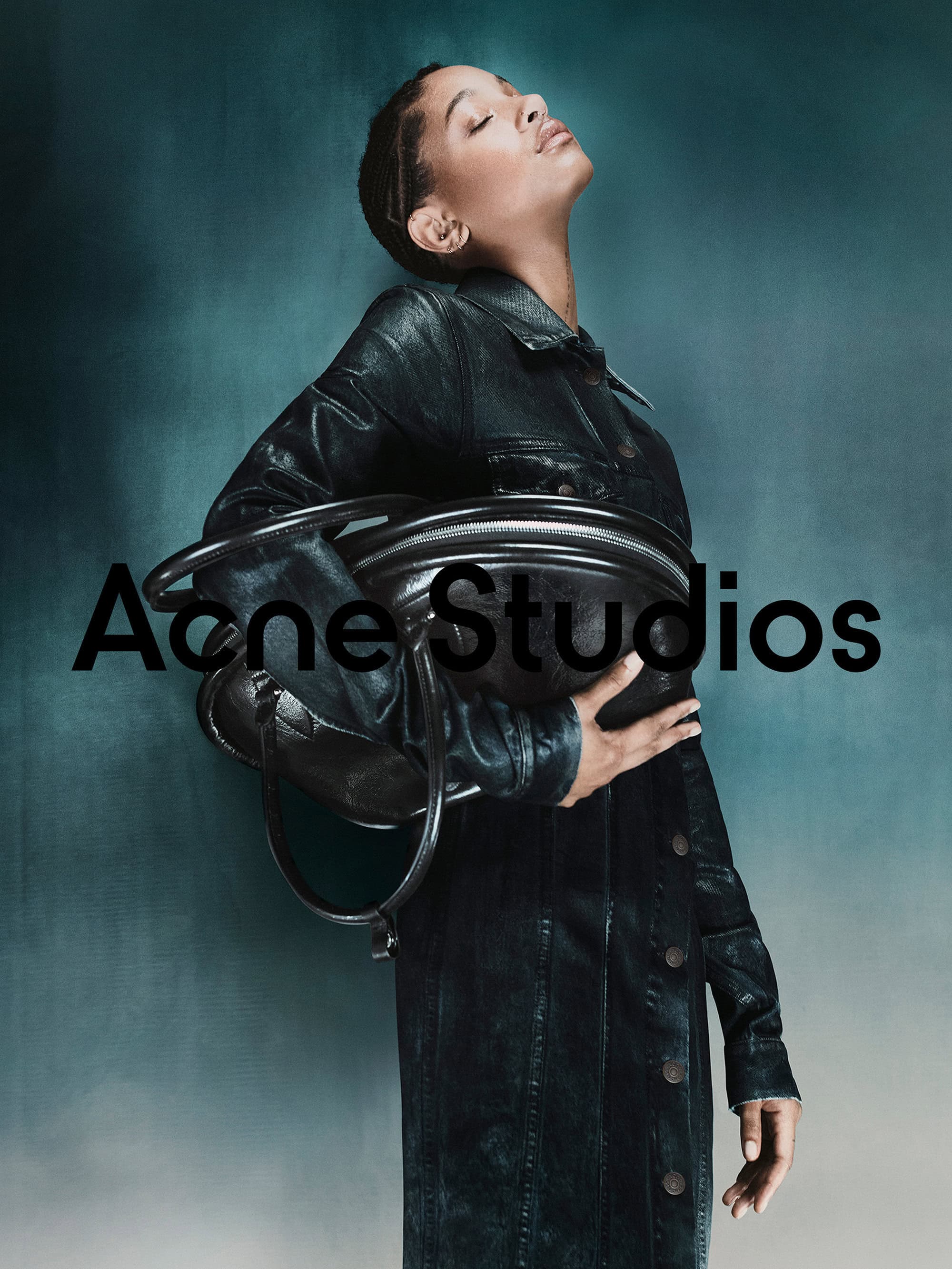 Willow Smith holding a dark leather bag wearing a matching dark coat against a teal gradient background