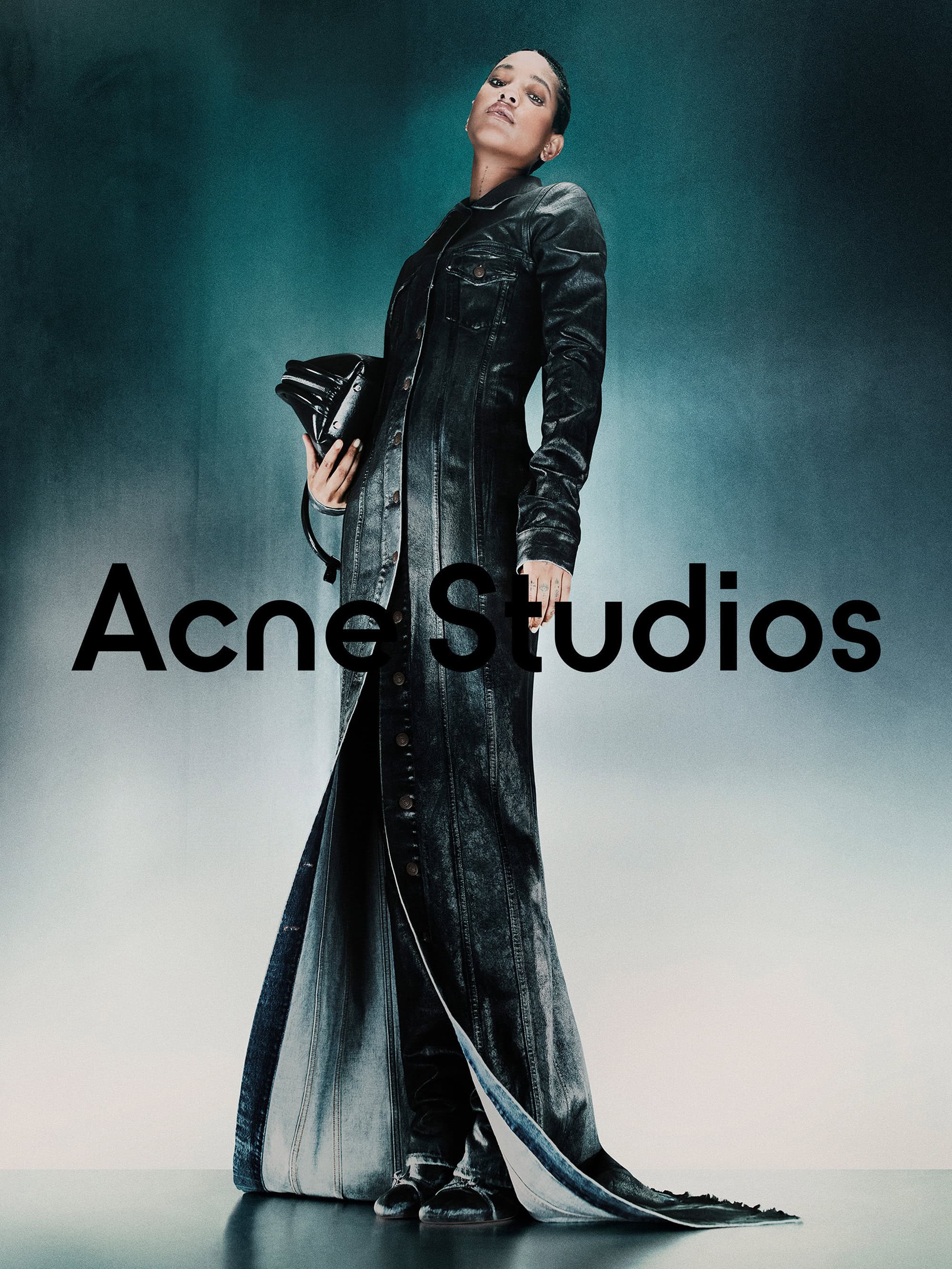 Model Willow Smith wearing a long dark denim coat holding a black handbag against a gradient backdrop
