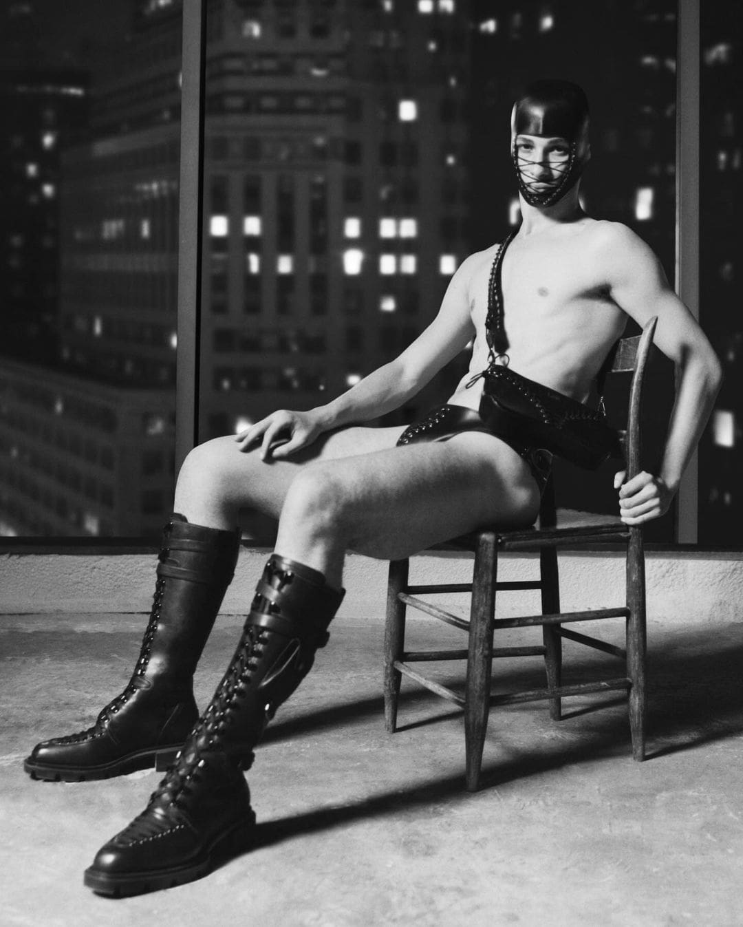 Model wearing a leather harness mask and tall boots sitting on a chair before an urban night window