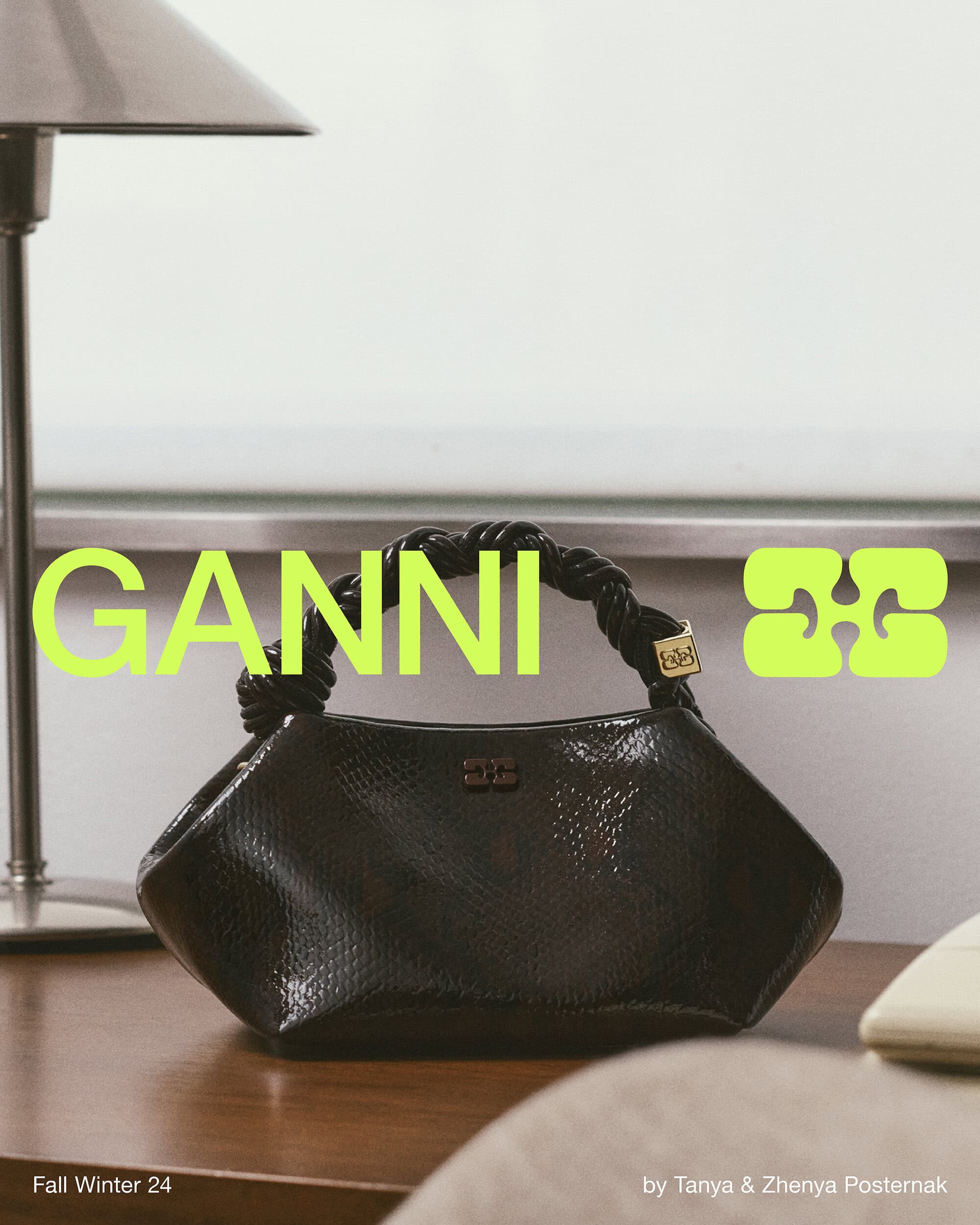 Black embossed leather handbag with a braided handle resting on a wooden desk next to a lamp for Ganni Fall 2024 campaign.