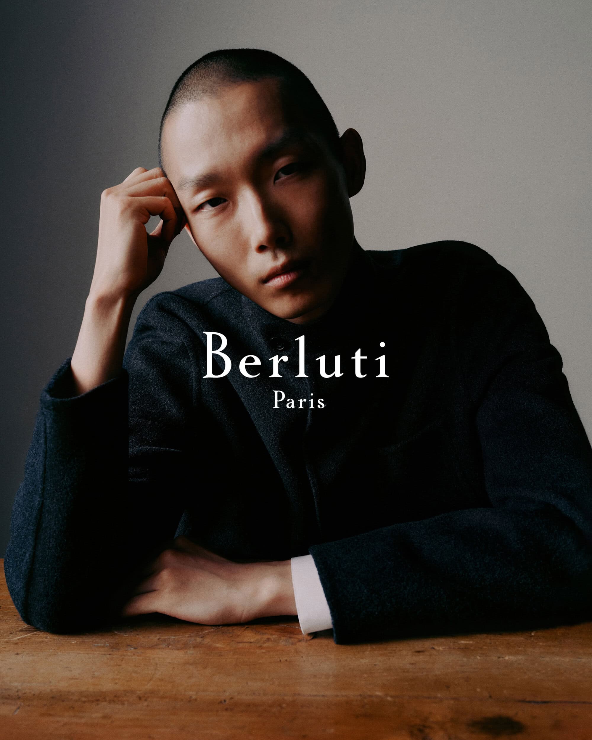 Close-up portrait of an East Asian male model wearing a dark jacket resting his head on his hand against a neutral studio background