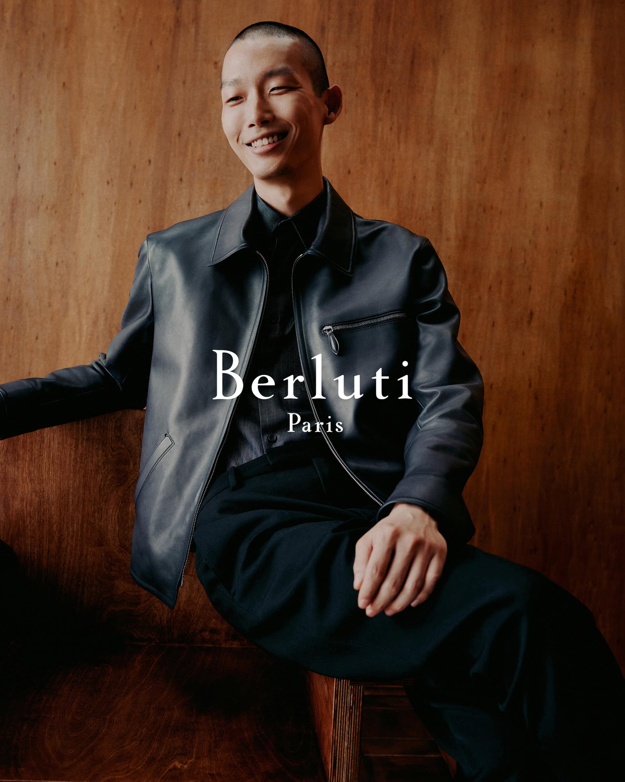 Model Xu Meen wearing a dark leather jacket seated against a warm wood paneled wall for Berluti Fall 2024 ad campaign.