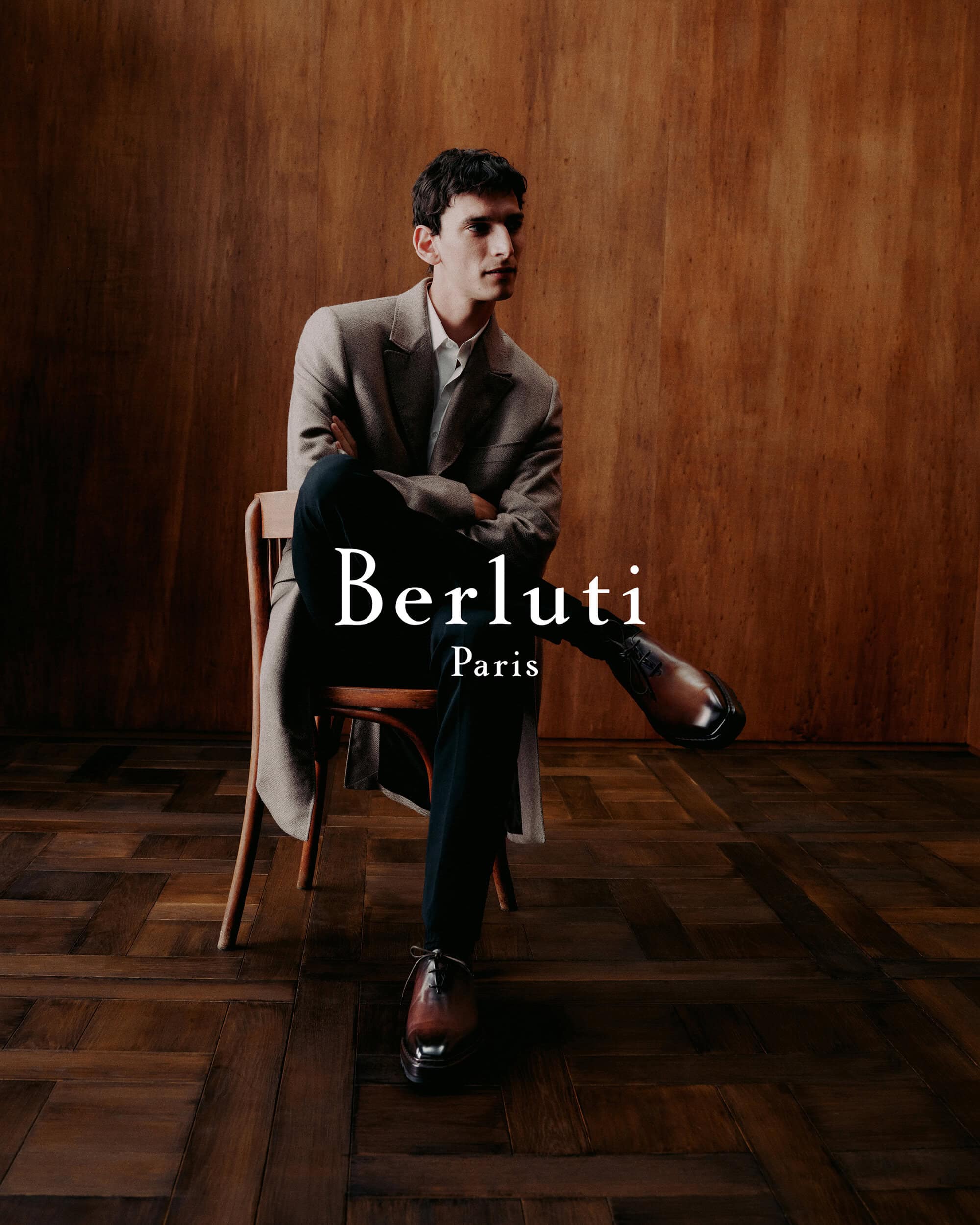 A man seated on a wooden chair wearing a camel coat and leather shoes against a dark wood panel wall.