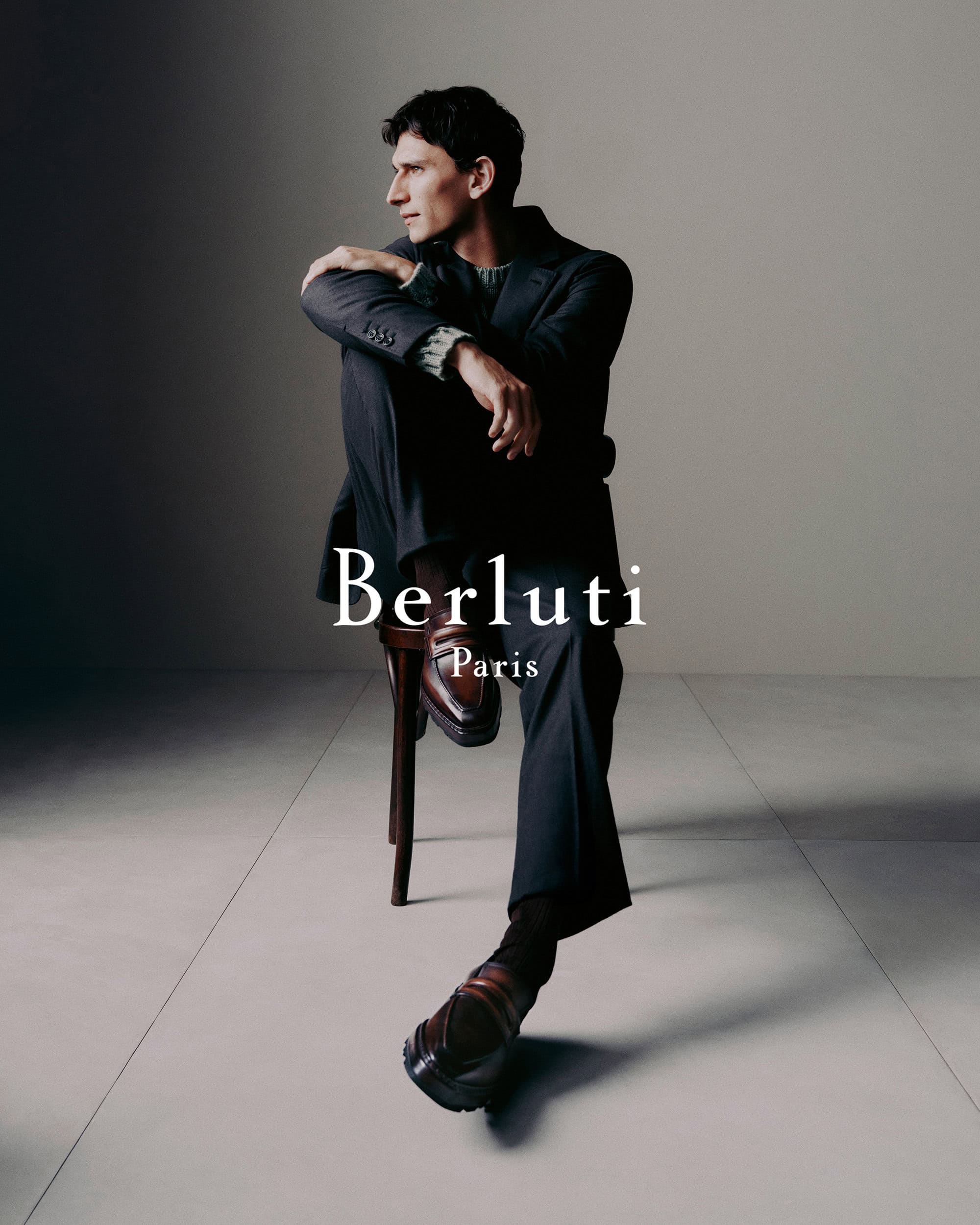 A man wearing a dark tailored suit and leather loafers seated on a wooden stool against a neutral studio backdrop.