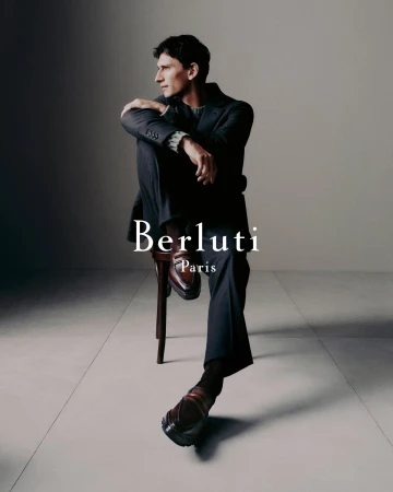 A man wearing a dark tailored suit and leather loafers seated on a wooden stool against a neutral studio backdrop.
