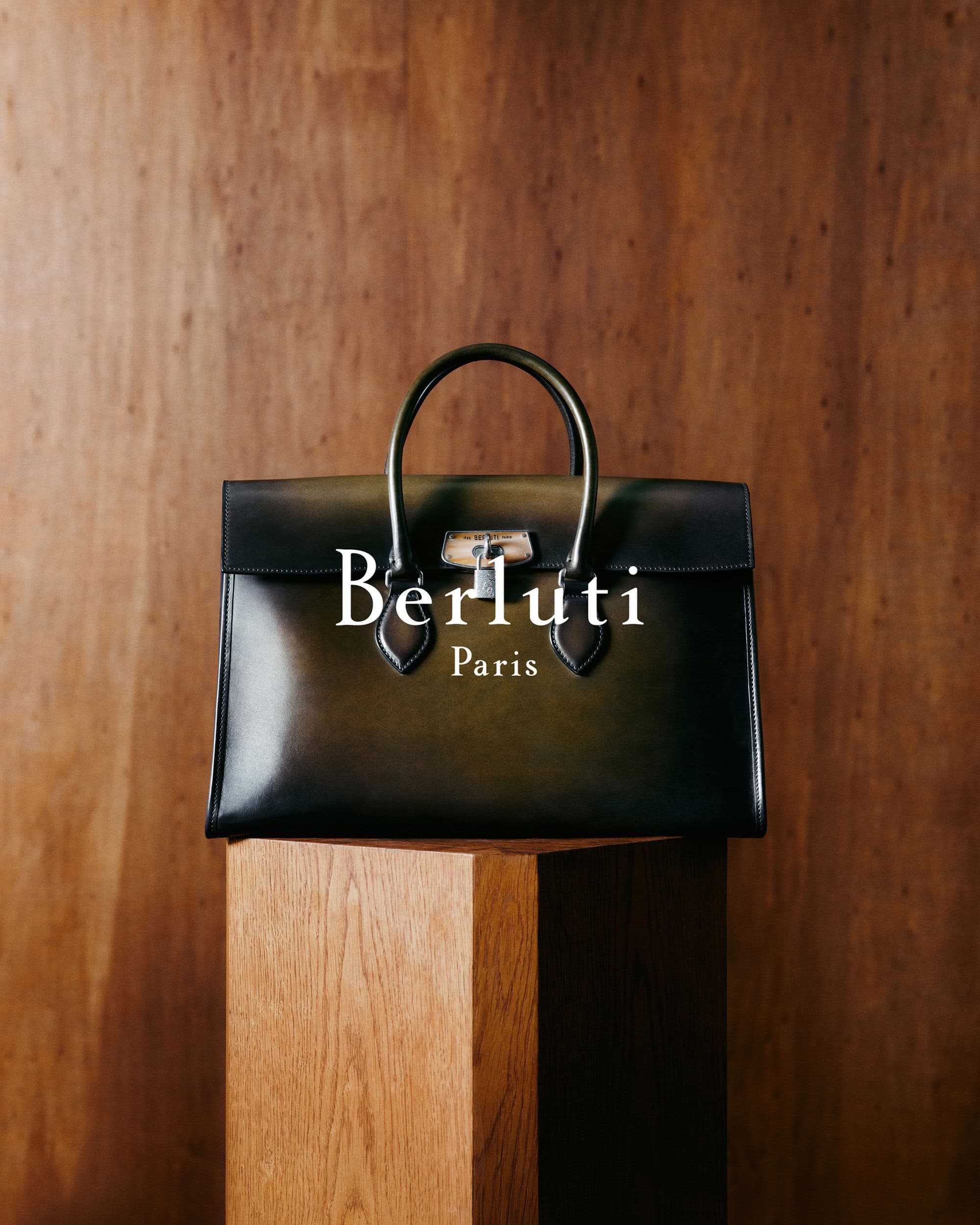 Berluti leather handbag displayed on a wooden pedestal against a dark wood panel background.