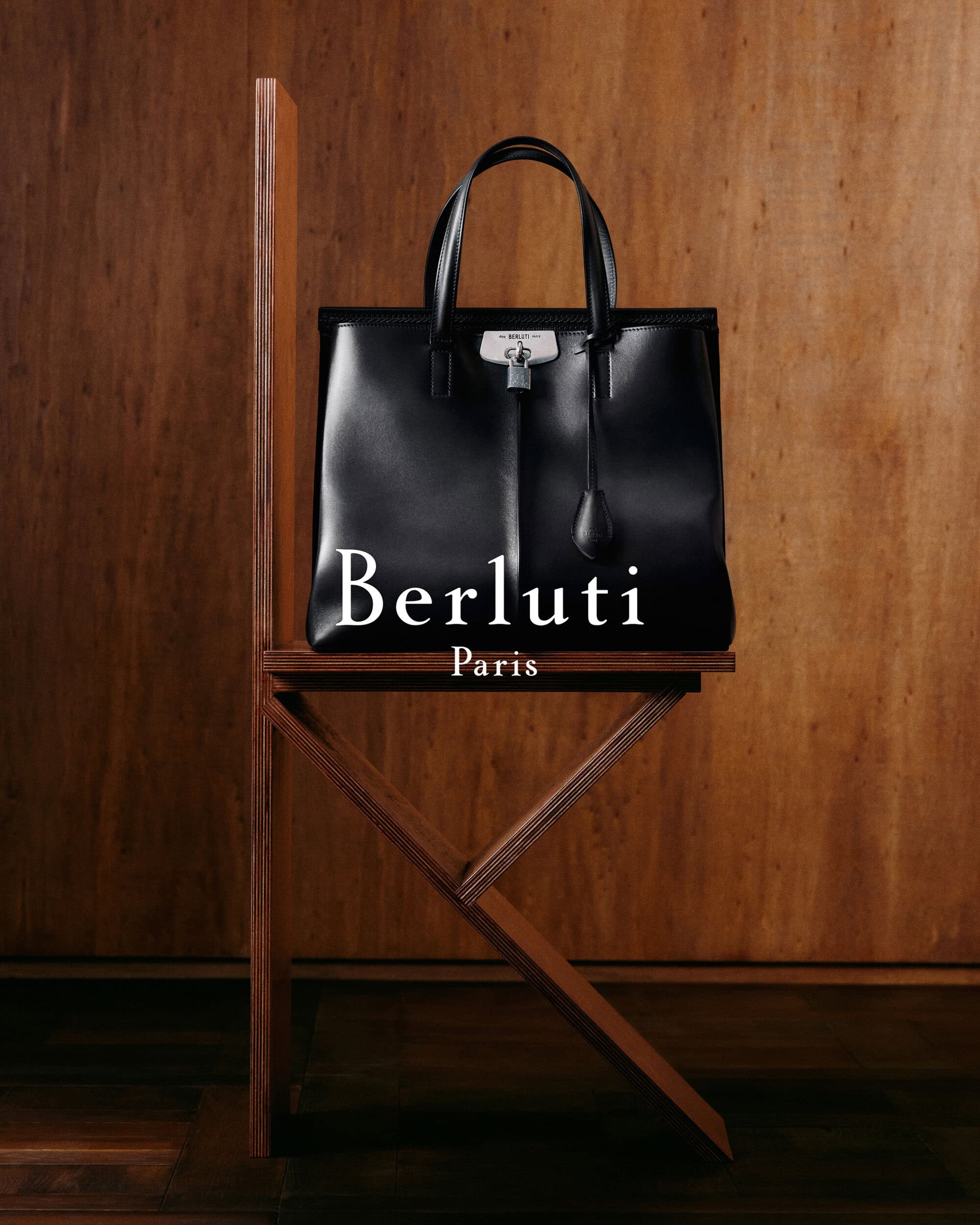 Black leather tote bag resting on a wooden sculptural chair against a wood paneled wall for Berluti Fall 2024 campaign