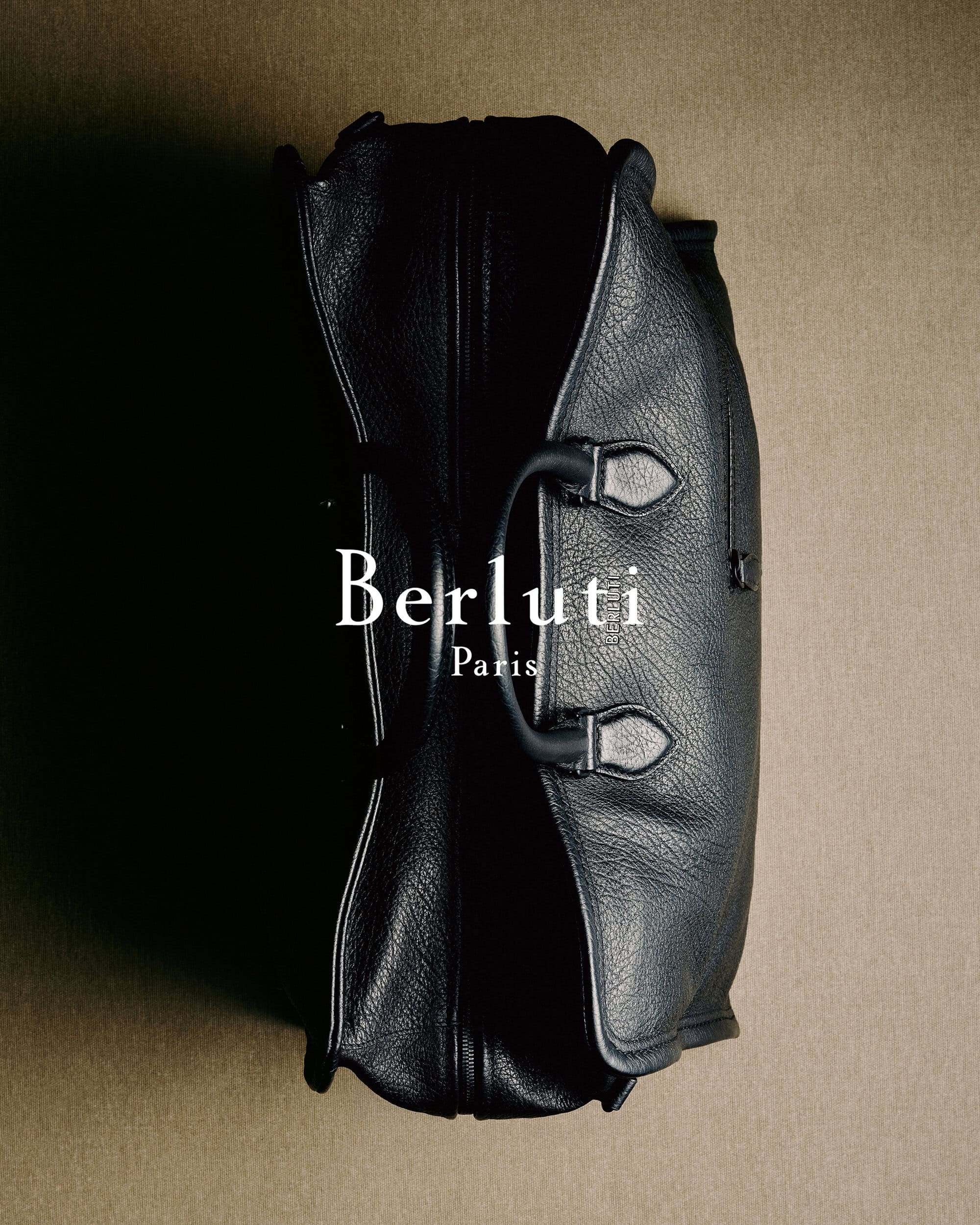 Black leather travel bag placed vertically on a textured neutral surface with visible brand typography