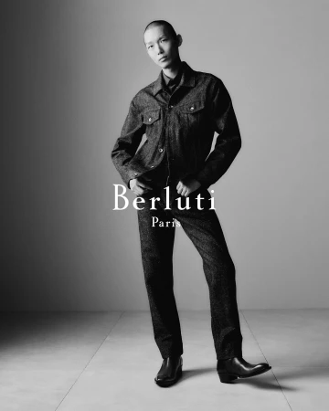 Monochrome fashion campaign image of a male model wearing a dark denim jacket and matching trousers against a grey seamless background