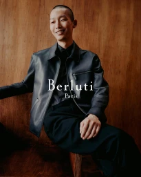 Model Xu Meen wearing a dark leather jacket seated against a warm wood paneled wall for Berluti Fall 2024 ad campaign.