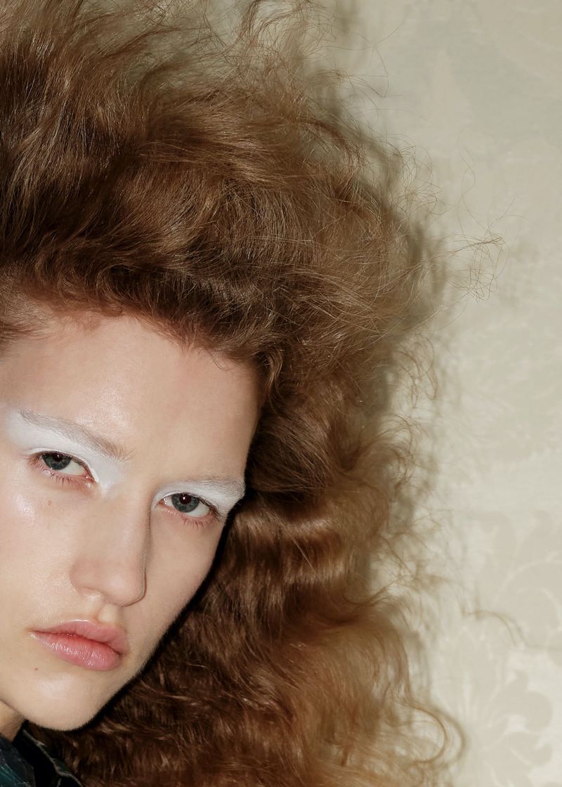 Close-up portrait of a young woman with large, wavy light brown hair and white eyebrow makeup posing against a neutral backdrop.