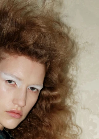 Close-up portrait of a young woman with large, wavy light brown hair and white eyebrow makeup posing against a neutral backdrop.
