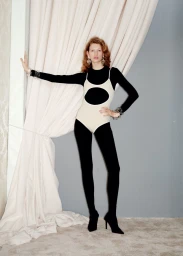 Model standing against heavy draped cream curtains wearing a black long-sleeve top under a cream cutout bodysuit with black tights.