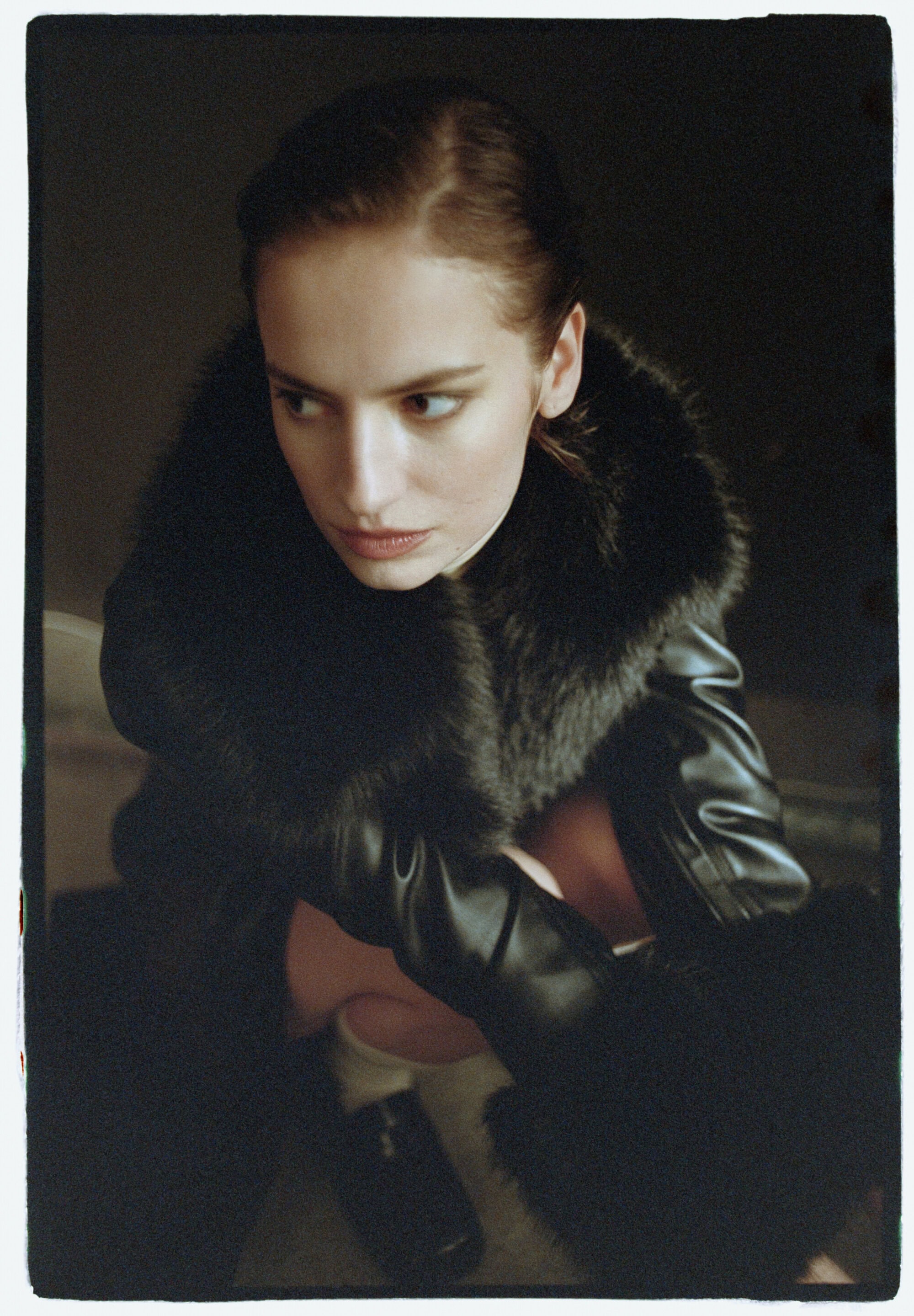 Woman in black faux fur jacket and dark loafers crouched against a dark background