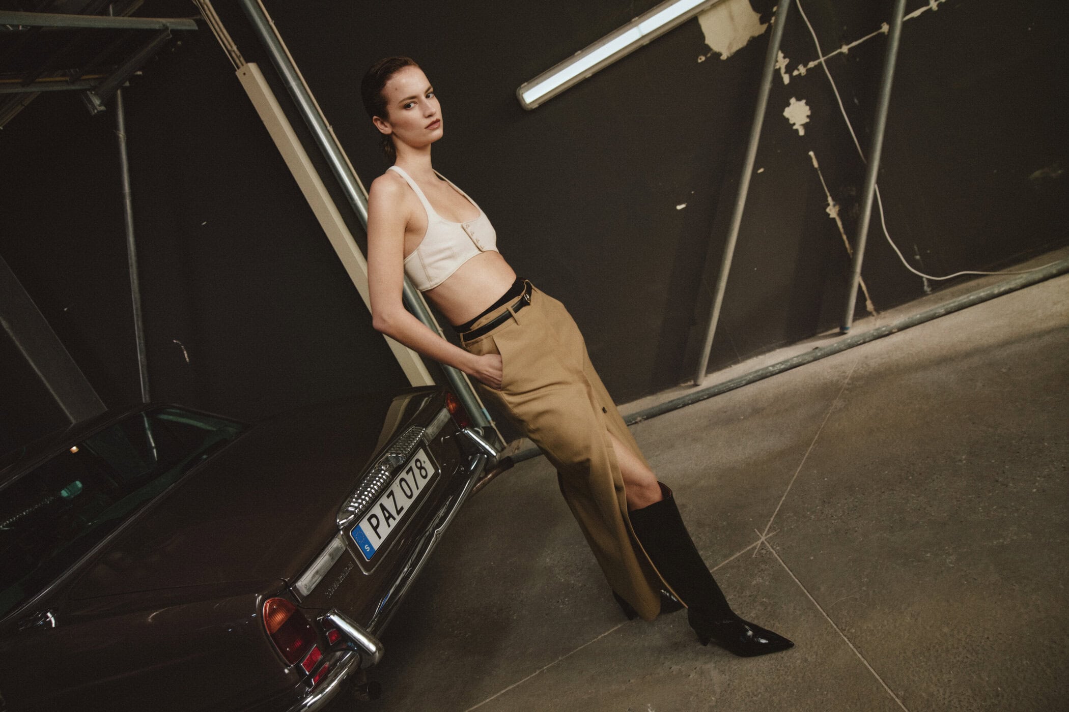 Model leaning against a vintage car wearing a white crop top and tan trousers in an industrial garage