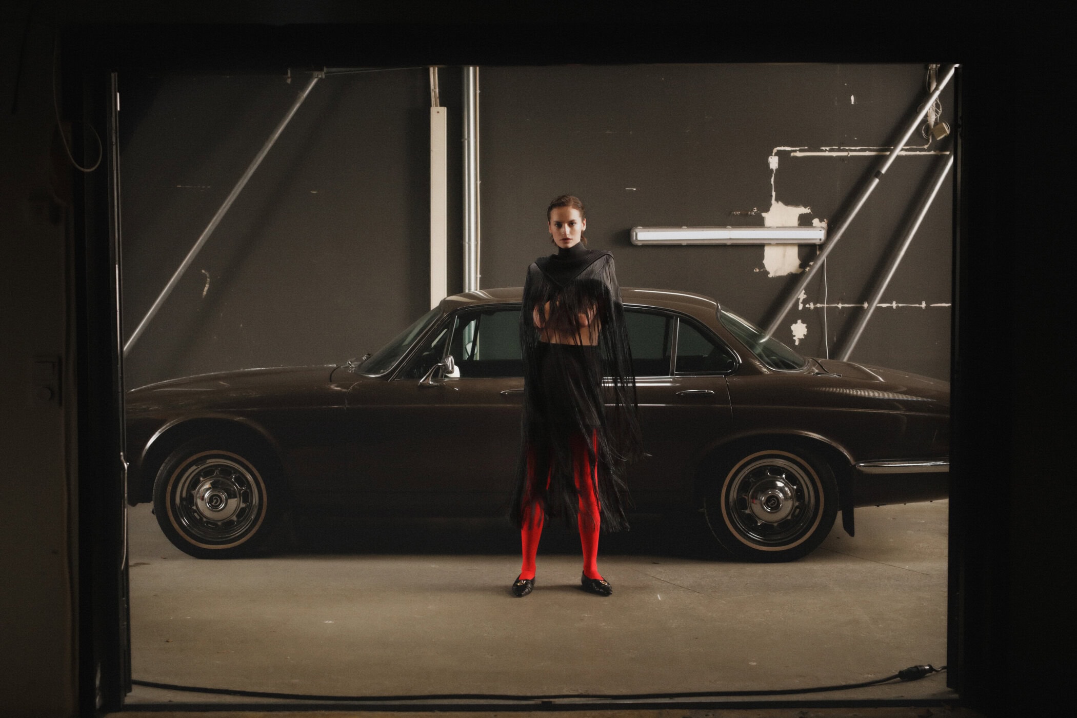 Model wearing black fringed garment and red tights standing in front of a vintage brown car in a garage