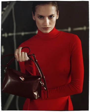 Woman in red turtleneck top holding a dark brown leather handbag against a dark background.