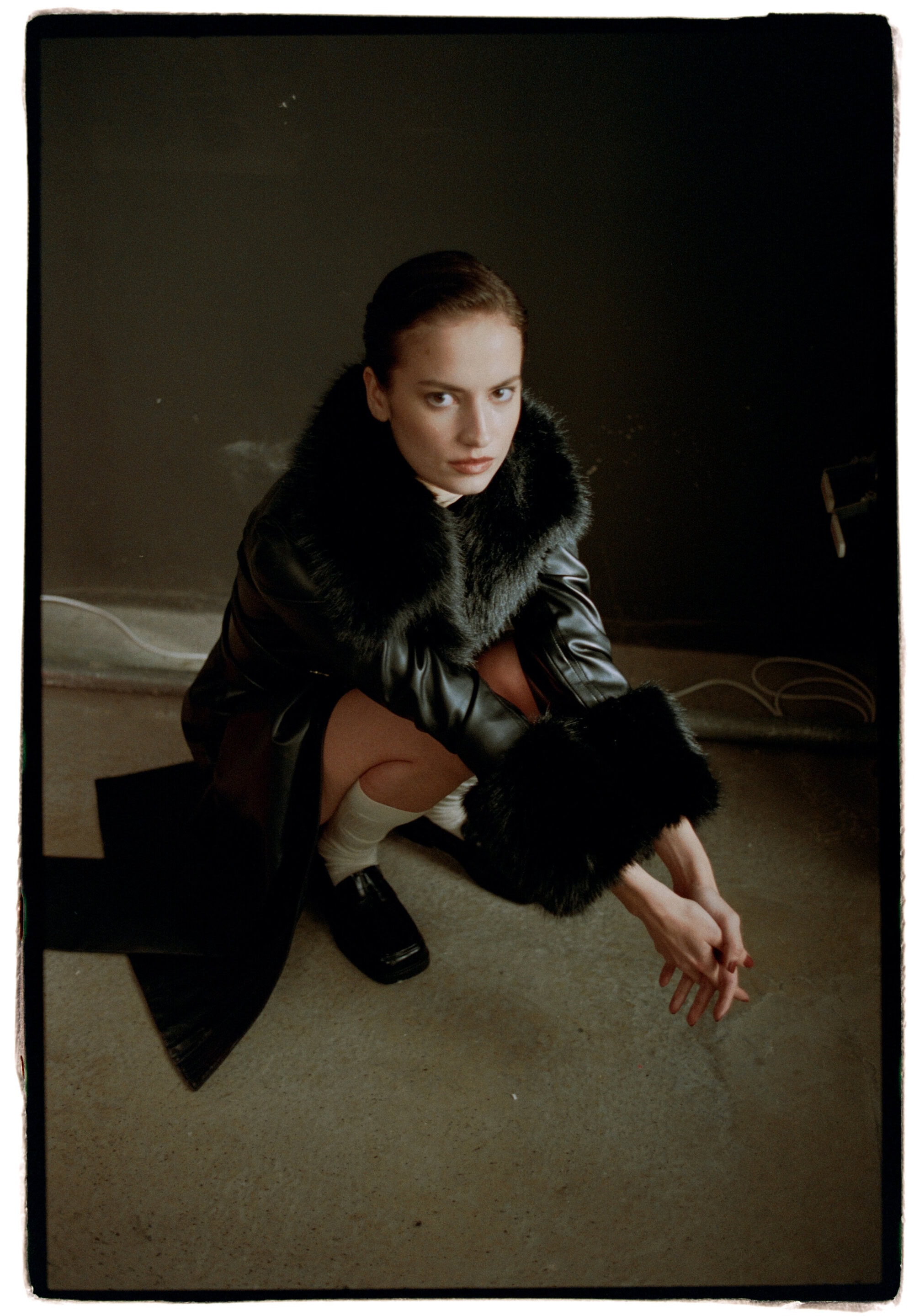 Woman crouching in a black faux fur trimmed coat and loafers against a dark studio background with black framing edges.