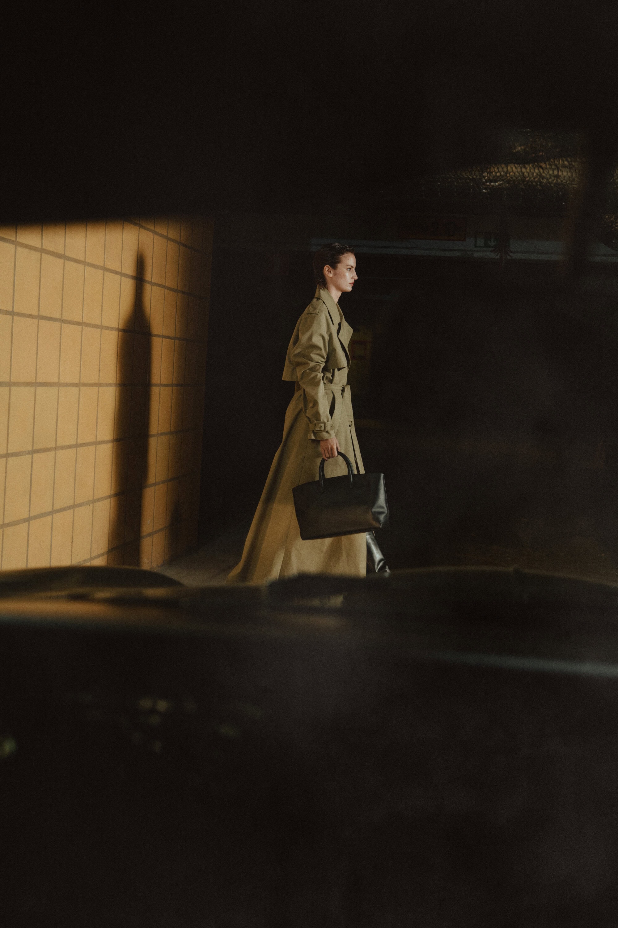 Model in a long beige trench coat holding a black handbag walking past a tiled wall for Flattered Fall 2024 campaign.