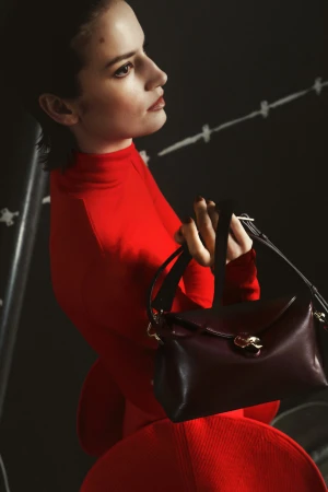 A woman in a vibrant red turtleneck top holding a dark brown leather handbag against a dark backdrop for the Flattered Fall 2024 campaign.