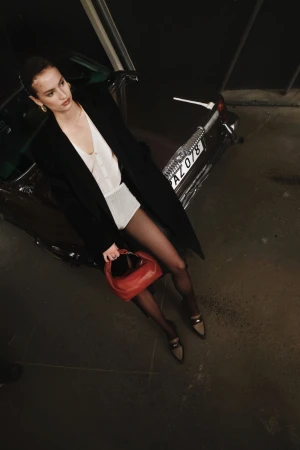 Woman in a black coat and white top holding a red handbag while leaning against a dark car for Flattered Fall 2024 Ad Campaign.