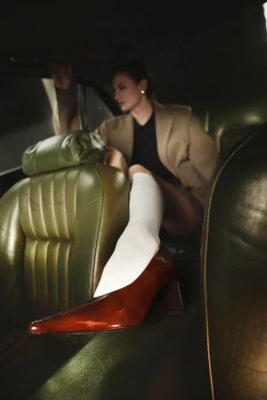 Woman in brown blazer sitting inside a car wearing white knee-high socks and shiny brown patent leather heels