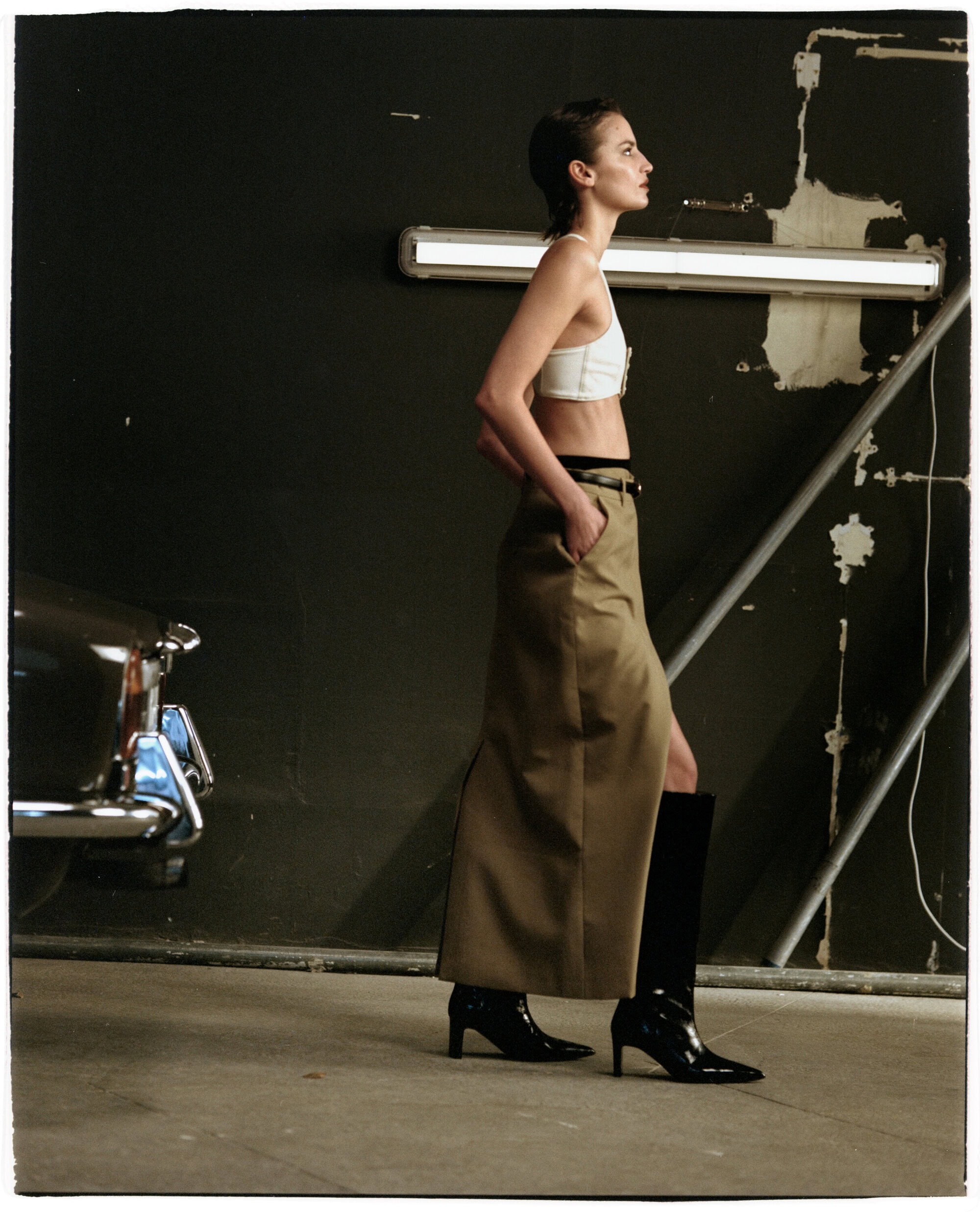 Model wearing a white crop top, beige skirt, and black boots posing in a garage next to a car for Flattered Fall 2024 campaign.