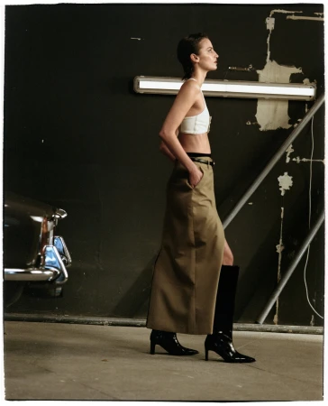 Model wearing a white crop top, beige skirt, and black boots posing in a garage next to a car for Flattered Fall 2024 campaign.
