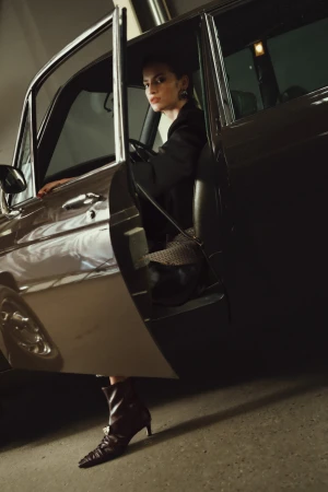 Woman in dark clothing sitting inside a vintage car with open door, wearing pointed boots.