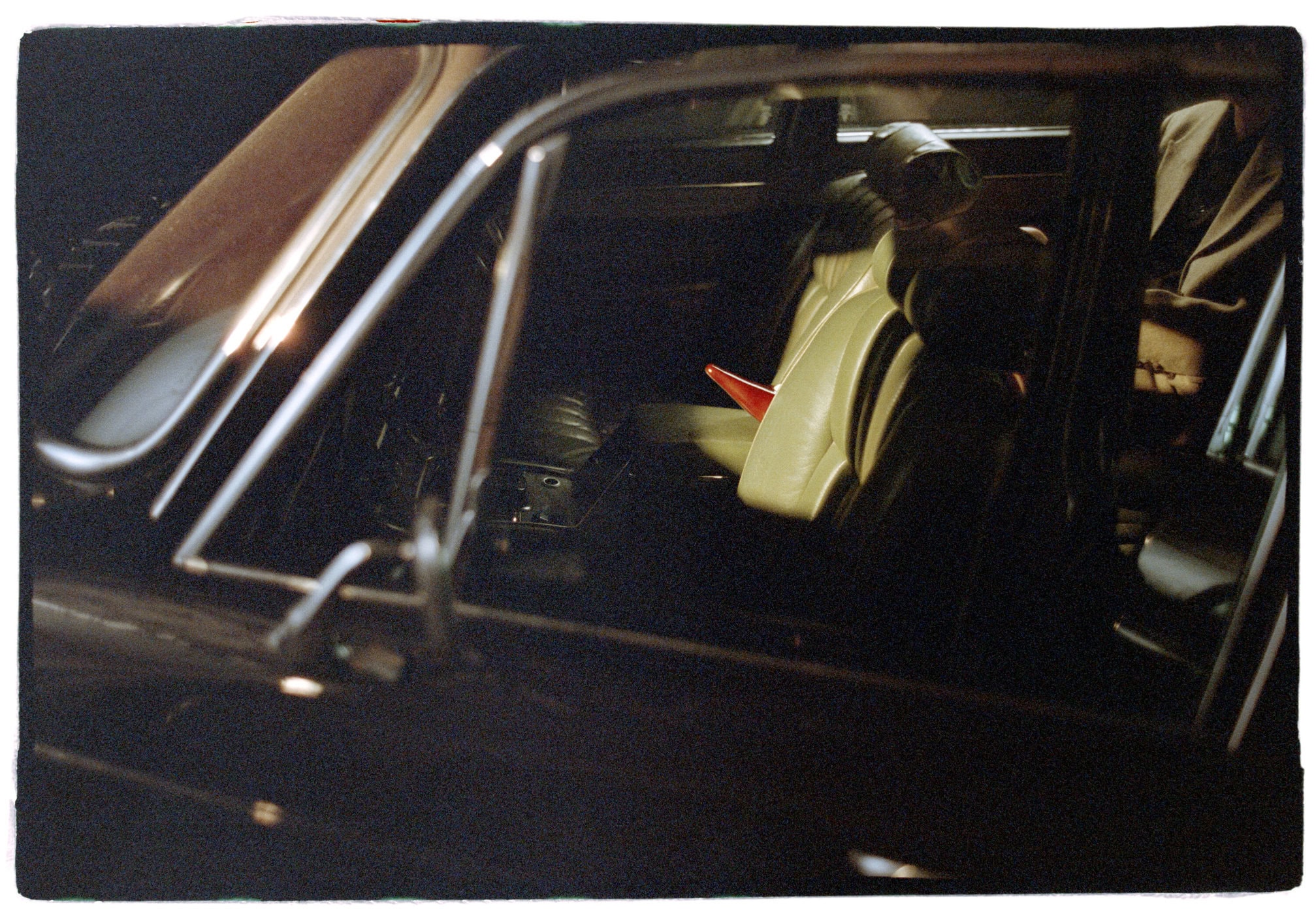 Interior of a dark vintage car at night showing light leather seats and a red shoe resting on the passenger seat.