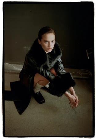 Woman crouching in a black faux fur trimmed coat and loafers against a dark studio background with black framing edges.