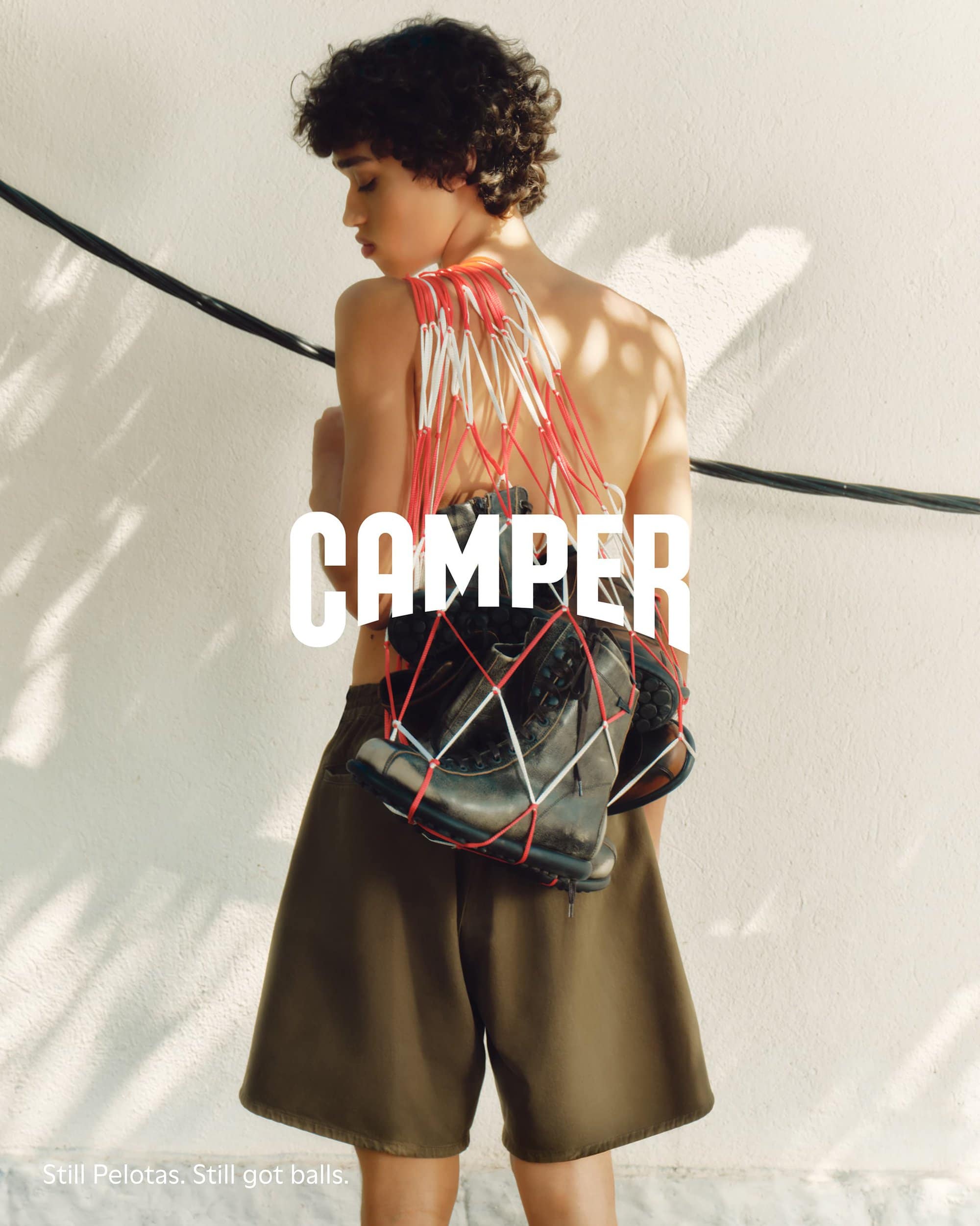 A shirtless model wearing olive shorts carries shoes in a red and white net bag for the Camper fall 2024 campaign.