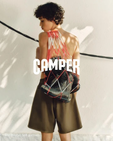 A shirtless model wearing olive shorts carries shoes in a red and white net bag for the Camper fall 2024 campaign.