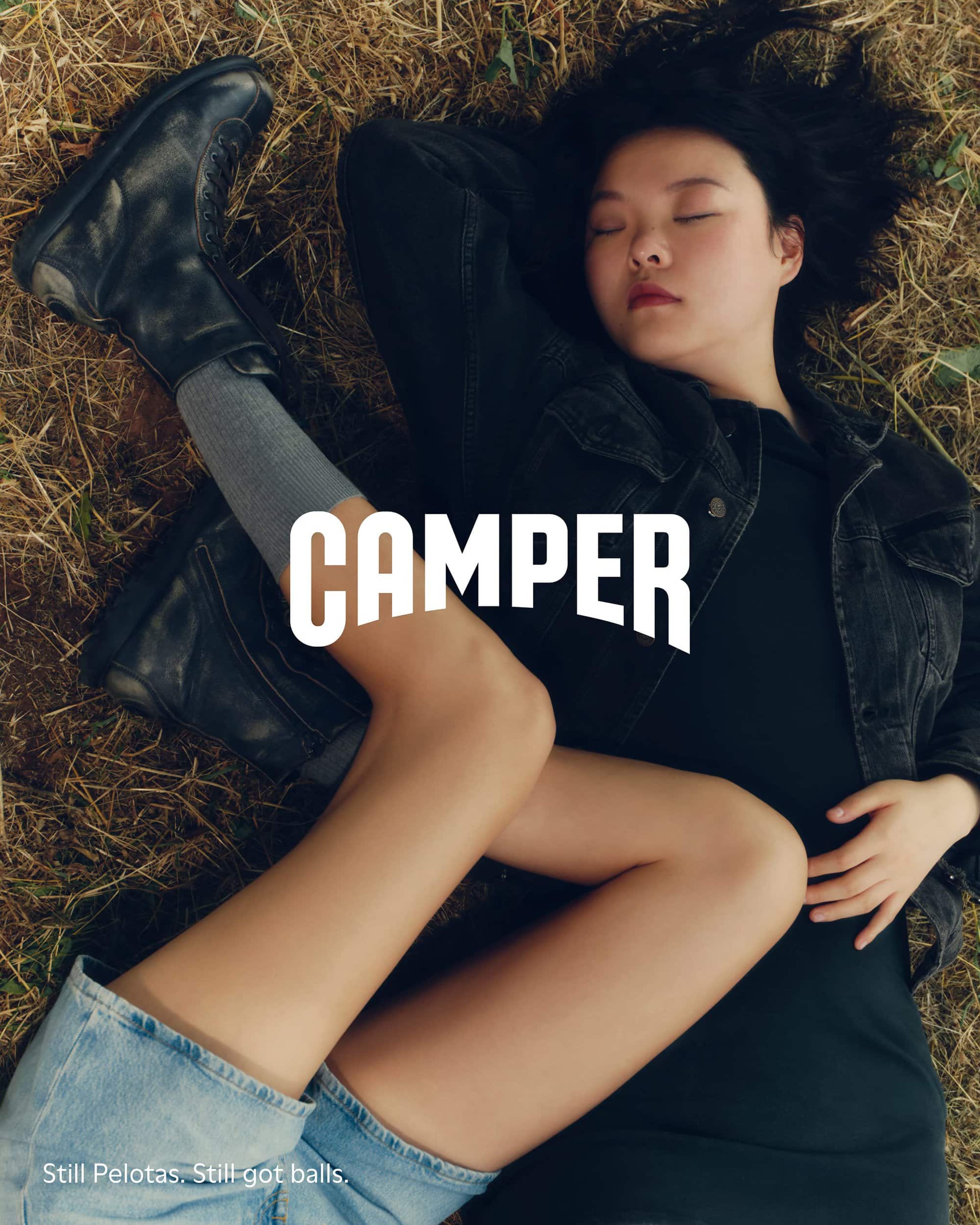 Model lying on dry grass wearing black jacket and denim shorts with black leather boots