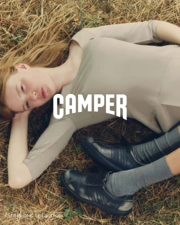 Model lying on dry grass wearing a beige top, grey socks, and dark sneakers for Camper Fall 2024 Ad Campaign