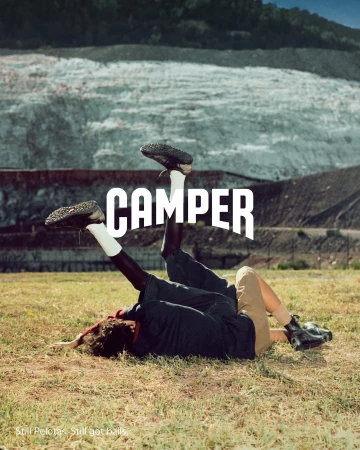 Two people lying on grass in an outdoor field with legs lifted up, featuring the Camper brand logo overlay.