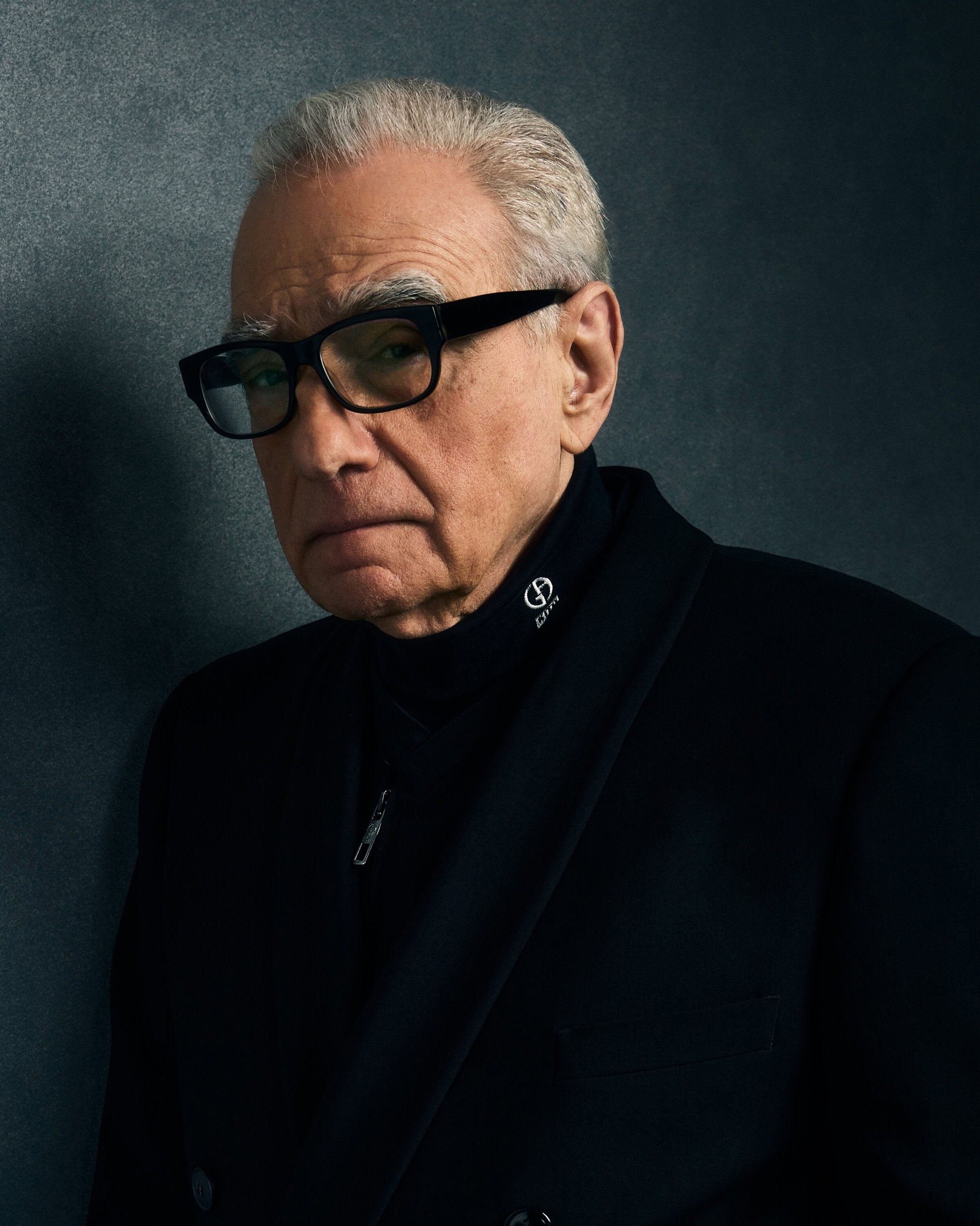 Martin Scorsese wearing black framed glasses and a dark coat against a dark textured studio backdrop