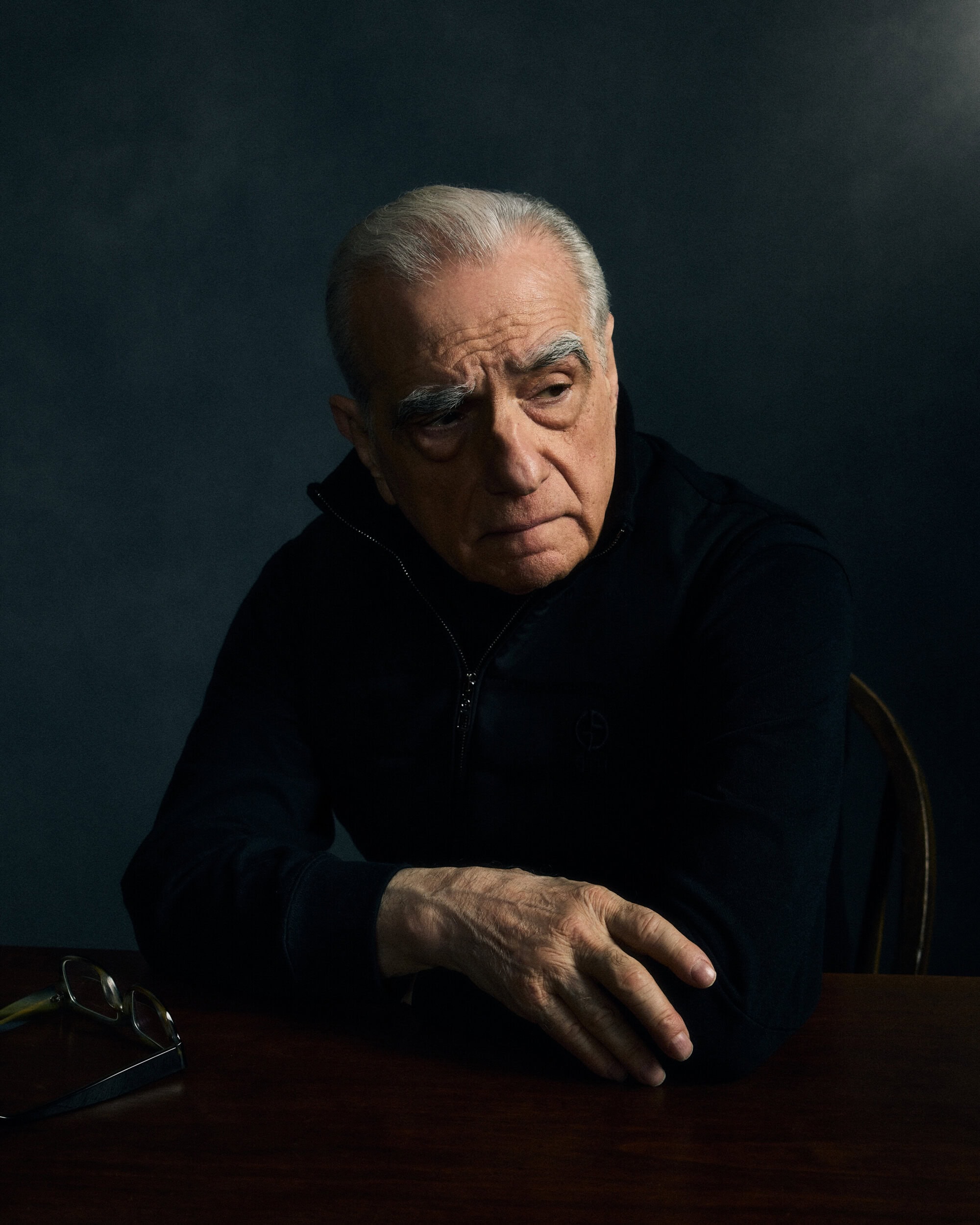 Portrait of Martin Scorsese wearing a dark zip-up jacket seated at a table against a dark textured backdrop