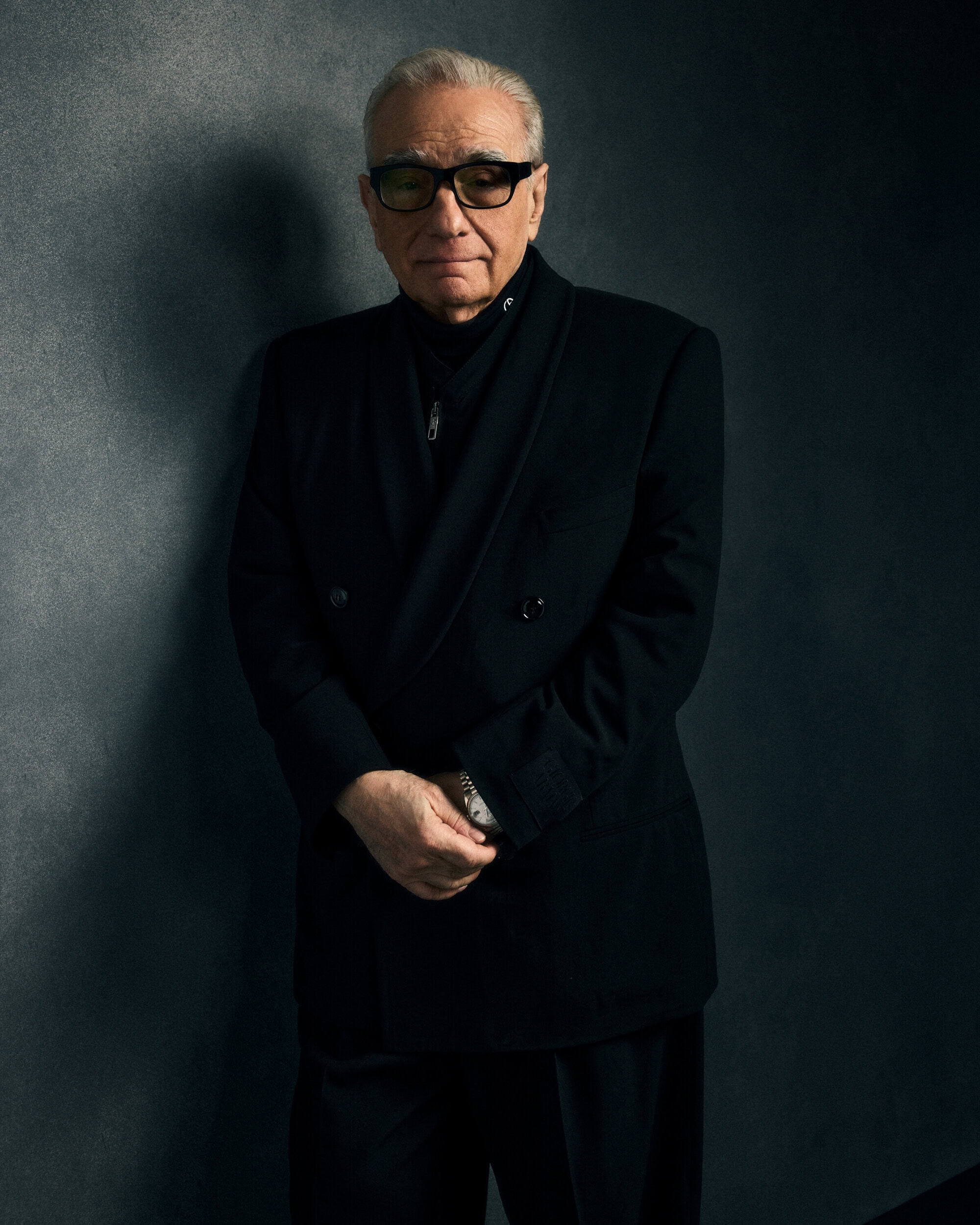 Martin Scorsese wearing a black double-breasted jacket and glasses standing against a dark textured wall