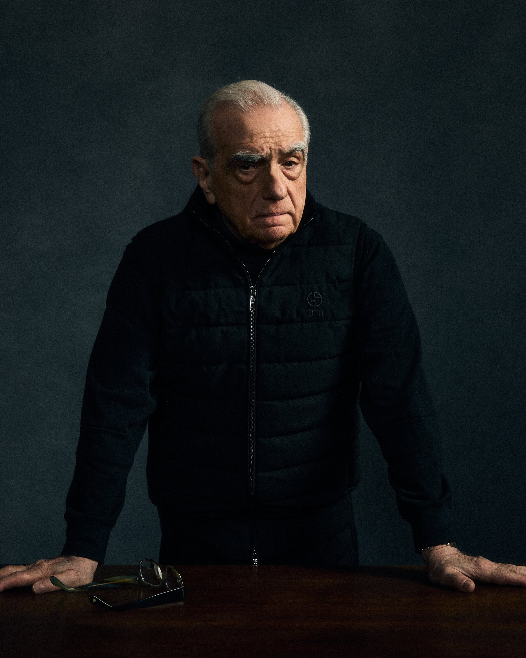 Martin Scorsese wearing a black quilted vest leaning on a wooden table against a dark textured background