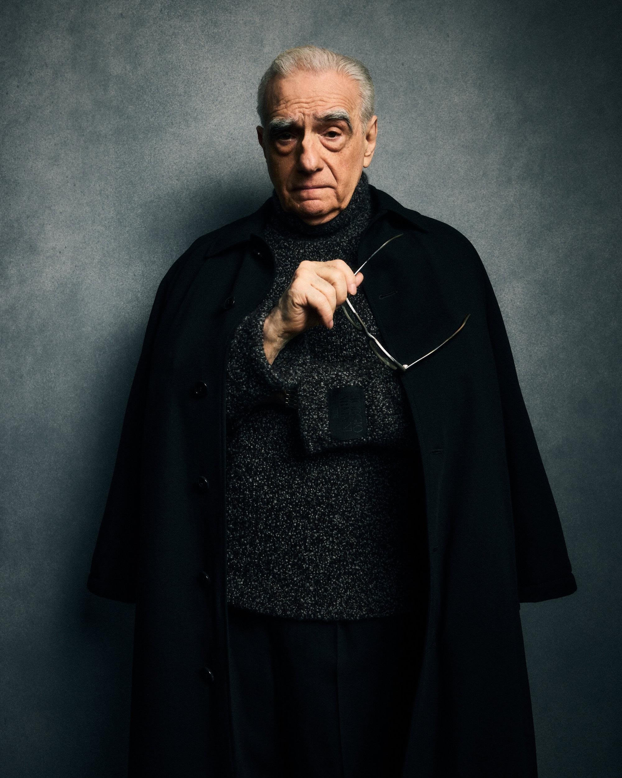 Portrait of Martin Scorsese wearing a dark coat and turtleneck sweater holding eyeglasses against a grey backdrop