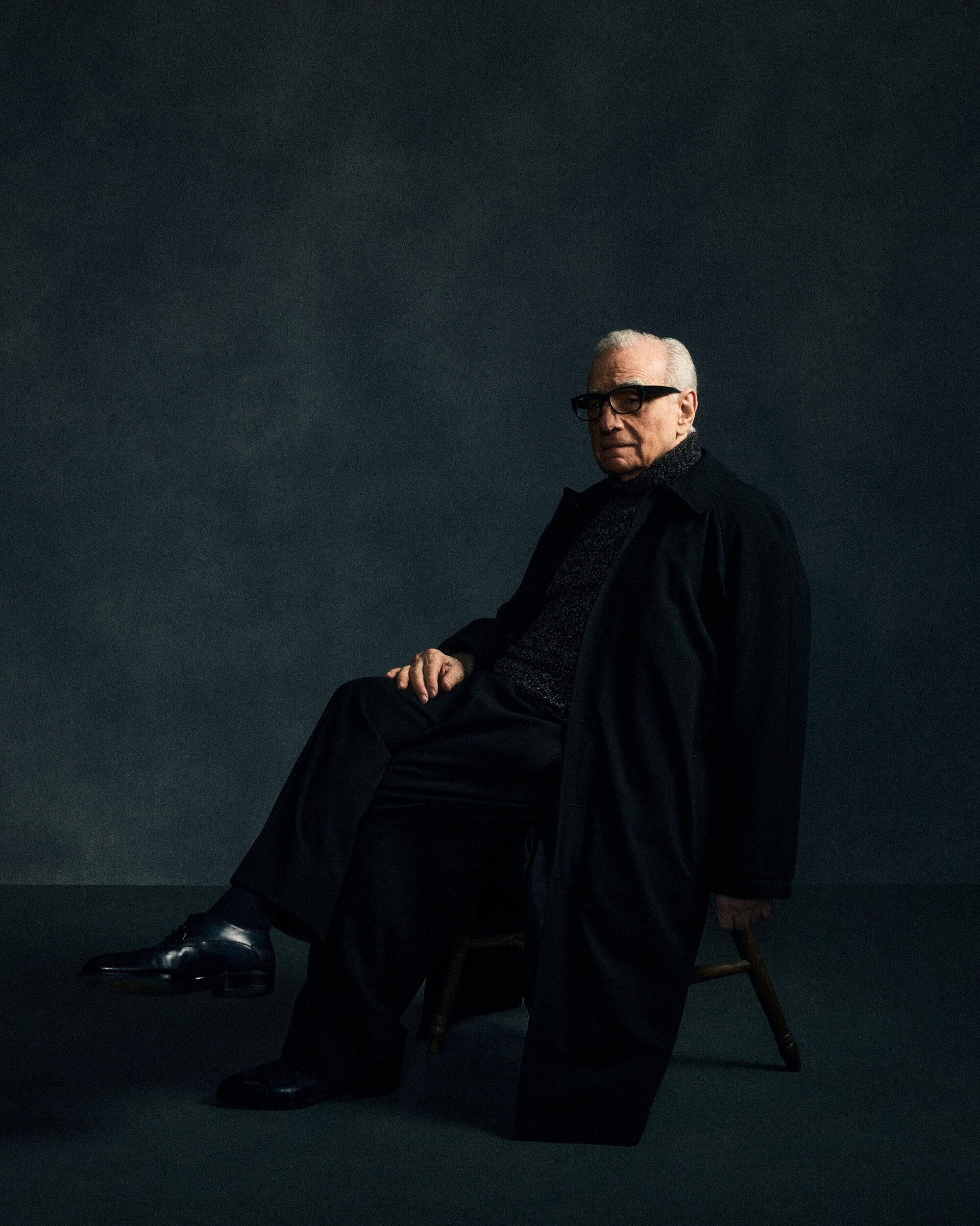 Martin Scorsese seated on a wooden chair against a dark textured studio backdrop wearing a black trench coat and glasses