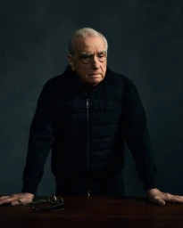 Martin Scorsese wearing a black quilted vest leaning on a wooden table against a dark textured background
