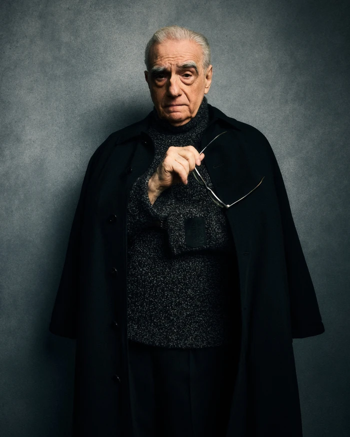 Portrait of Martin Scorsese wearing a dark coat and turtleneck sweater holding eyeglasses against a grey backdrop