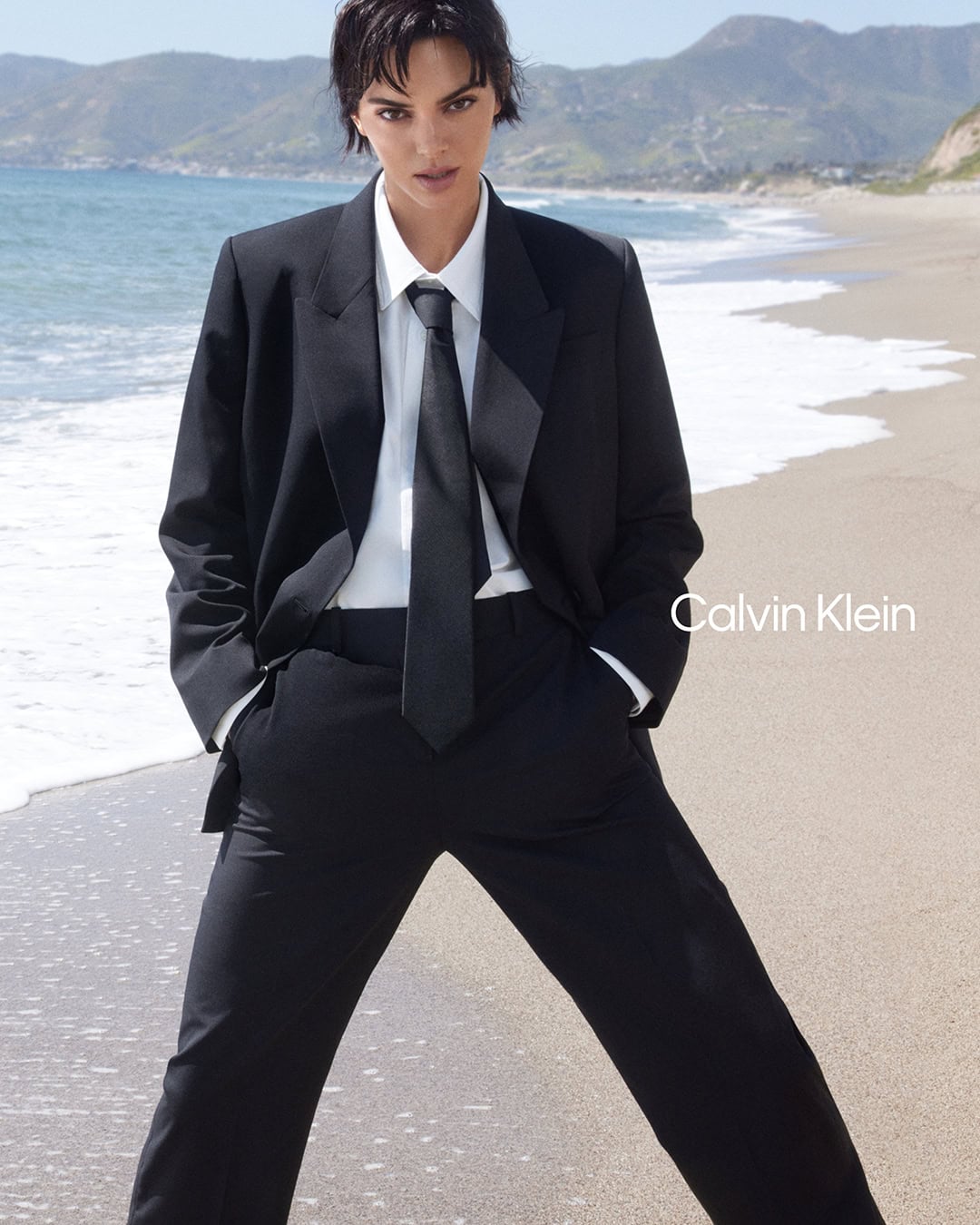 Model Kendall Jenner wearing a black suit and tie on a sandy beach for Calvin Klein Fall 2024 ad campaign.